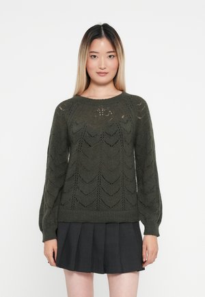 Grüner gestrickter Pullover mit Chevron-Muster, rundem Halsausschnitt und langen Ballonärmeln. Kombiniert mit einem gefalteten schwarzen Rock.