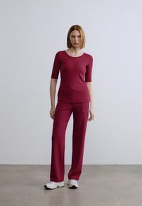 Μπουφάν σε μπορντό χρώμα με κοντά μανίκια, συνδυασμένο με παντελόνια φαρδιάς γραμμής σε ίδιο χρώμα και πλευρικές τσέπες, φορεμένο με λευκά αθλητικά παπούτσια. Ομαλή υφή.