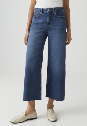 Frau trägt blaue, weite, kurz geschnittene Jeans mit rohen Säumen, weißes ärmelloses Top und cremefarbene Slip-On-Schuhe, steht vor schlichtem Hintergrund.