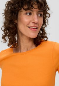 Jasnopomarańczowa, prążkowana koszulka z okrągłym dekoltem. Posiada dopasowany krój i krótkie rękawy. Gładka faktura, minimalne detale szwów.