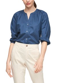 Mujer vestida con una camisa azul abotonada de mangas tres cuartos y pantalones crema con un sutil patrón floral, con las manos relajadas a los lados.