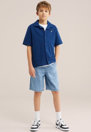 Dječak stoji obučen u plavu košulju s kratkim rukavima s gumbima, svijetle traper kratke hlače, bijele čarape i crno-bijele tenisice na jednobojnoj pozadini.