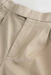 Beige Hochbundhose mit glatter Textur, vorne mit Falten und breitem Bund. Keine sichtbaren Beschläge oder Muster.