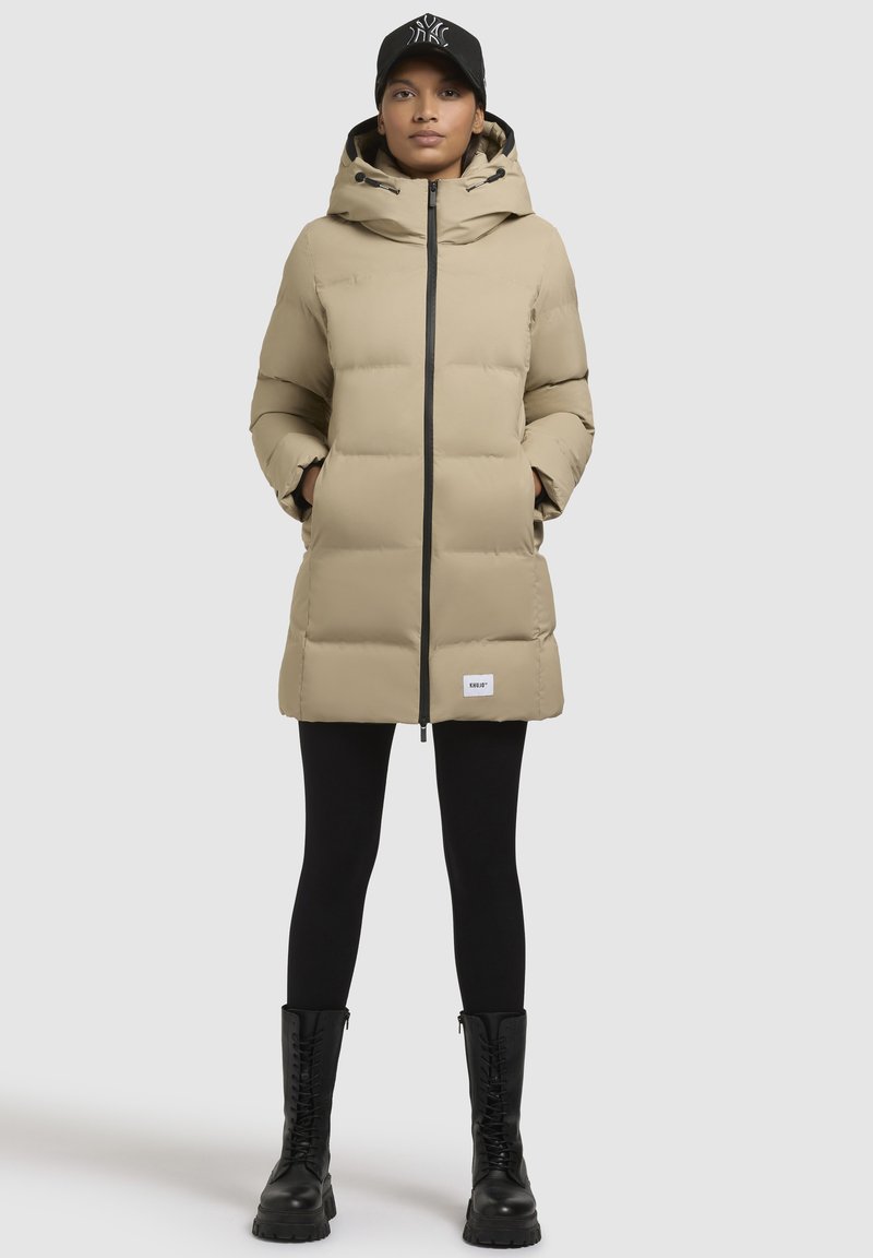 Beige pufferjas met een capuchon, voorzien van een rits aan de voorkant, zijzakken en zwarte accenten. Draagt over zwarte leggings en veterschoenen.