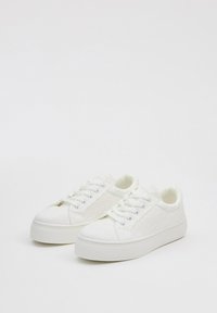 Zapatillas blancas con material superior texturizado, punta redonda, suela plana y cierre con cordones. Presentan un sutil detalle de patrón en la superficie.