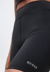 Κοντινό πλάνο μαύρων αθλητικών σορτς με ψηλή μέση και μικρό λευκό λογότυπο "GUESS" στον αριστερό μηρό.