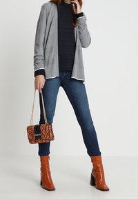 Grå cardigan med svarta detaljer, marinblå topp, mörka denimjeans och handväska i ormskinnsprint i orange. Glansiga orange höga stövlar med fyrkantiga tår.