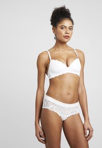 Witte kanten bh met geschulpte randen en verstelbare banden, gecombineerd met sheer witte string met bloemkanten afgewerkt.