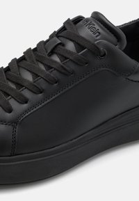 Černá kožená teniska s kulatou špičkou, texturovaným povrchem, černými tkaničkami a polstrovaným límcem. Obsahuje hladkou gumovou podrážku.