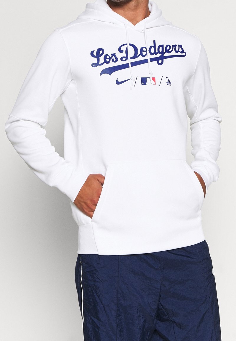 Personne portant un sweat à capuche blanc Los Dodgers avec une poche avant et un pantalon bleu marine, les mains partiellement dans les poches du sweat.