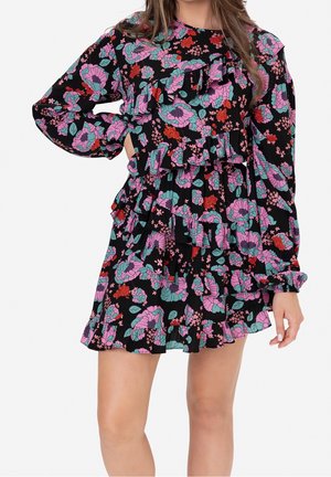 Mujer con un vestido negro con estampado floral con volantes rosas, rojos y azul verdoso, mangas largas y una falda con capas, de pie con una mano en la cadera.