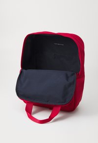 Sac à dos rouge avec un rabat bleu foncé, fabriqué en tissu durable. Il dispose d'une poignée supérieure et d'un intérieur spacieux pour le rangement.