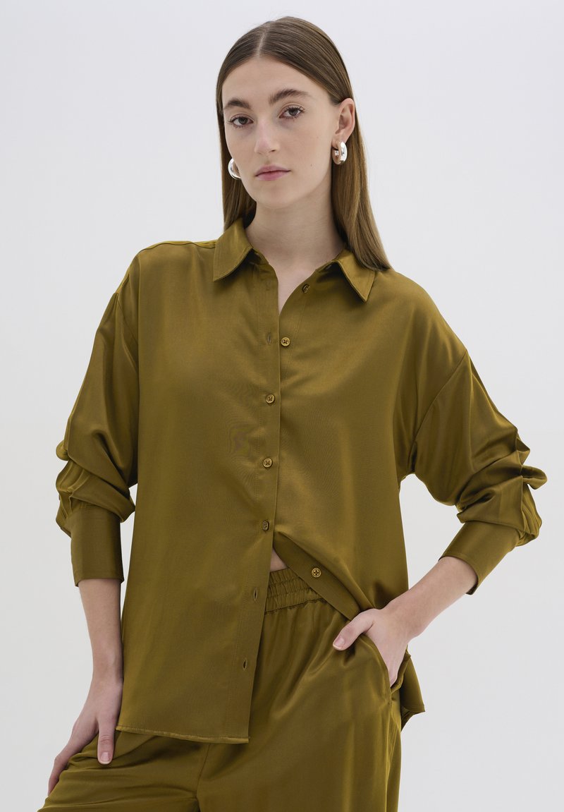 Camicia in satin verde oliva con bottoni frontali, colletto e maniche lunghe. Presenta una texture morbida e una vestibilità rilassata, abbinata a pantaloni coordinati.