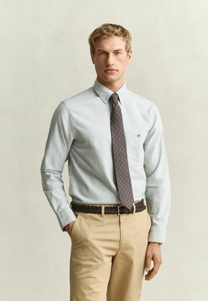 Homme portant une chemise bleu clair à boutons, une cravate à motifs, un pantalon beige et une ceinture sombre tressée, debout avec une main dans la poche.