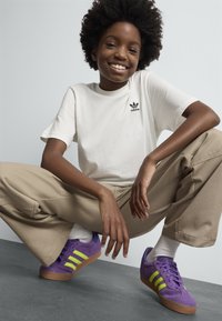 Mosolygó, göndör hajú személy fehér Adidas pólót, bézs nadrágot, fehér zoknit és lila Adidas cipőt visel neon zöld csíkokkal, guggoló helyzetben.