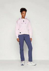 Šviesiai rožinė zip-up gobtuvė su raišteliais, derinama su tamsiai mėlynomis kelnėmis ir baltomis sportinėmis bateliais su oranžiniais akcentais. Paprastas ir funkcionalus dizainas.