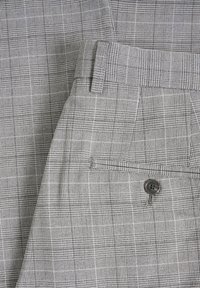 Grå rutigt tyg med små rutor och linjer, med en knapp och fickdetalj på den vikt delen.