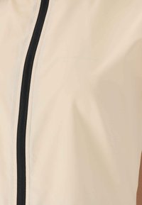 Vaalean beige kevyt takki, jossa on musta vetoketju edessä, sileä kangas ja minimalistinen muotoilu ilman lisäkuvioita tai koristeita.