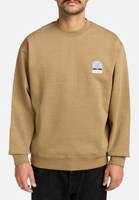 Sweatshirt marron à manches longues, avec des poignets côtelés et un col rond. Présente un petit logo de palmier brodé sur la poitrine.