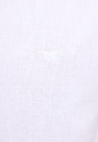 Tessuto di lino bianco con una texture sottile, caratterizzato da un piccolo dettaglio ricamato in bianco che somiglia a un cavallo sulla superficie.