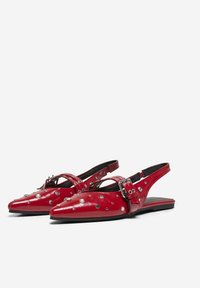 Zapatos rojos tipo slingback con punta afilada, material de cuero brillante, acentos de tachuelas y detalle de hebilla ajustable en la correa.