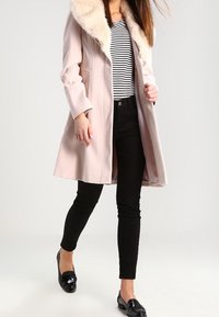Ljusrosa ullkappa med fuskpälskrage, bärs över en randig skjorta och svarta byxor. Svarta blanka loafers fulländar outfiten.