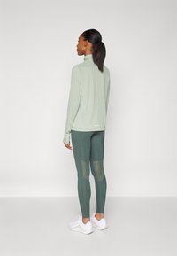 Maglietta a maniche lunghe verde chiaro abbinata a leggings verde scuro con pannelli in rete, completata da scarpe da ginnastica bianche.