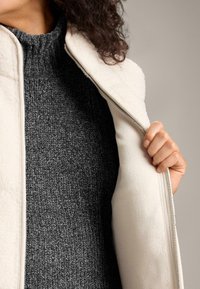 Person trägt einen dunkelgrauen Strickpullover und hält eine cremefarbene Fleecejacke mit Reißverschluss vor einem schlichten Hintergrund geöffnet.