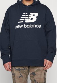 Henkilö, jolla on yllään laivastonsininen New Balance -huppari, jossa on etutasku, sekä maastokuvioiset cargo-housut, seisomassa vaaleaa taustaa vasten.