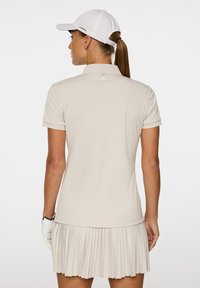 Beżowa koszulka polo golfowa z plisowaną spódnicą. Krótkie rękawy, miękka tkanina i mały detal z logo z tyłu. Rękawiczki noszone na lewej ręce.