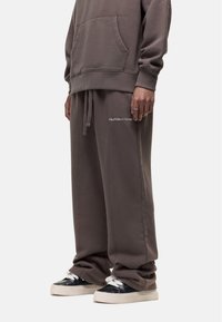 Loose-fit, brown sweatpants with waistband drawstring, side pockets, and embroidered details. Paired with black sneakers featuring white laces.