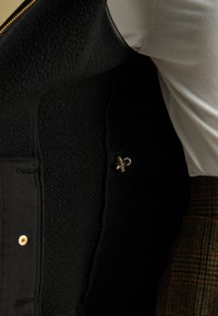 Čierny fleecový vest s hladkou textúrou, s detailom zlatého kovu. Obsahuje zips a prešité švy.