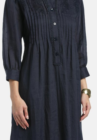 Tmavomodré šaty s dlhými rukávmi, predným zapínaním na gombíky, vertikálnymi záhybmi na hrudi, kvetinovou výšivkou a zlatým náramkom na pravom zápästí.