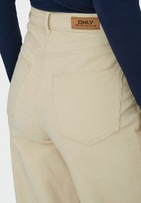 Pantalon en velours côtelé beige avec taille haute, doté de deux poches arrière et d'une étiquette en cuir pour le détail. La texture est côtelée et douce.