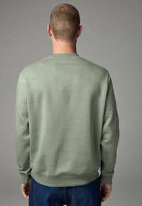 Hele roheline kets, ümarate kaelustega, ribakudumistega varrukate ja äärega, sileda tekstuuriga ning minimalistikku disainiga. Vaade tagant.