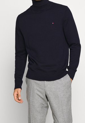 Marineblauer Rollkragenpullover aus weichem Strickmaterial, mit einem kleinen roten und weißen Logo auf der Brust. Kombiniert mit grauen strukturierten Hosen.