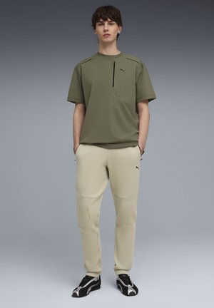 Olivgrünes Kurzarmhemd mit einer Brusttasche, kombiniert mit beigen Jogginghosen mit Seitentaschen und schwarzen Akzenten. Schwarze Schuhe.