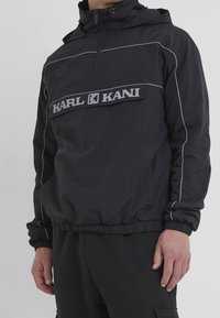 Black hooded jacket with front zip, elastic cuffs and hem, white piping on sleeves, and "KARL KANI" logo across the chest pocket.