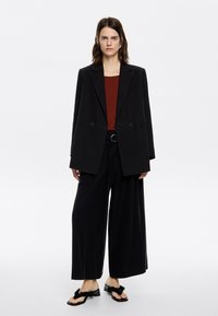 Zwarte oversized blazer met twee knopen aan de voorkant, gecombineerd met wijde zwarte broek en zwarte sandalen met decoratieve franjes.