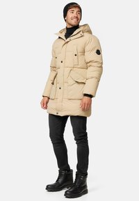 Beige színű pufferszövet kabát, kapucnival, fekete gombokkal és zsebekkel. Fekete, slim-fit farmernadrággal és fekete fűzős csizmával párosítva.