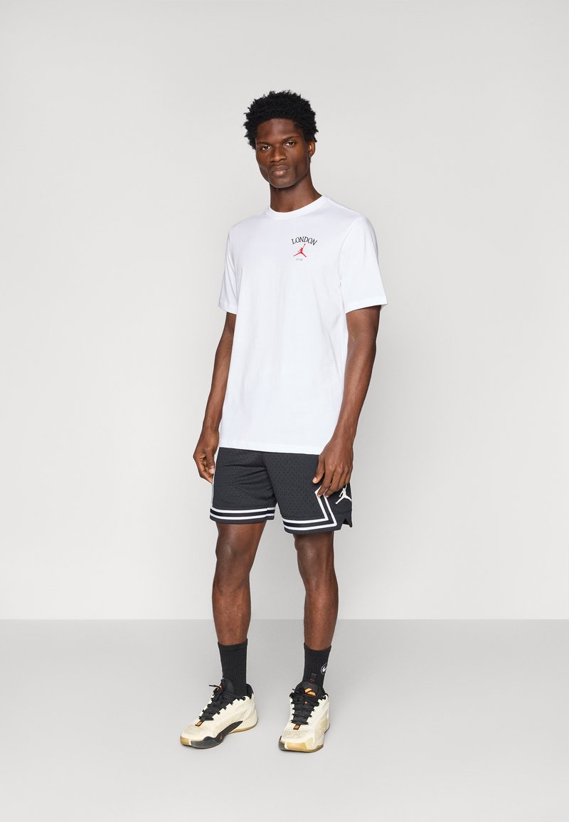 Un homme debout portant un t-shirt blanc "London", un short de basketball noir avec des rayures blanches, des chaussettes noires et des baskets blanches sur fond uni.