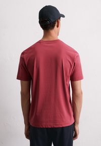 Rode katoenen T-shirt met korte mouwen en een ronde hals, gezien vanachter. Zwarte pet met verstelbare sluiting complementeert de outfit.