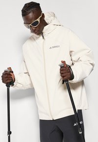 Beige Terrex jacket features a zip closure, adjustable hood, and elastic cuffs, paired with gray pants and trekking poles.