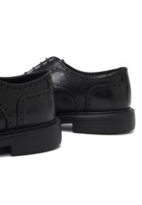 Zwarte leren schoenen met brogue-details, met een gladde afwerking en een ronde neus, rustend op een zwarte rubberen zool met horizontale groeven.