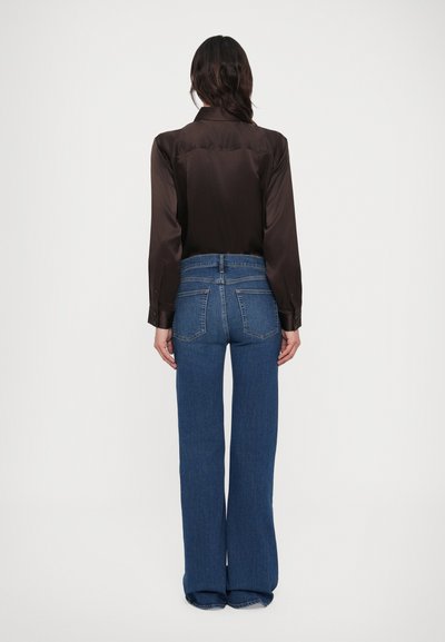 Nő háttal áll, sötétbarna hosszú ujjú ingben, melyet magas derekú, széles szárú kék farmernadrágba tűrt, fehér háttér előtt.