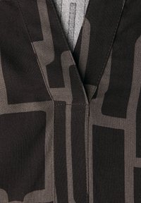 Tessuto marrone con motivi geometrici grigi, caratterizzato da una scollatura a V e dettagli di cucitura visibili. Vista ravvicinata del materiale e del design.