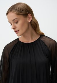 Zwarte geplisseerde jurk met transparante mouwen en een ronde hals. Glad materiaal met een vloeiende design, waardoor een lichte silhouet ontstaat.