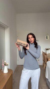 Scatola di cartone marrone con testo nero "zalando", tenuta da una persona in un dolcevita grigio. Mobili in legno chiaro e decorazioni neutre sullo sfondo.