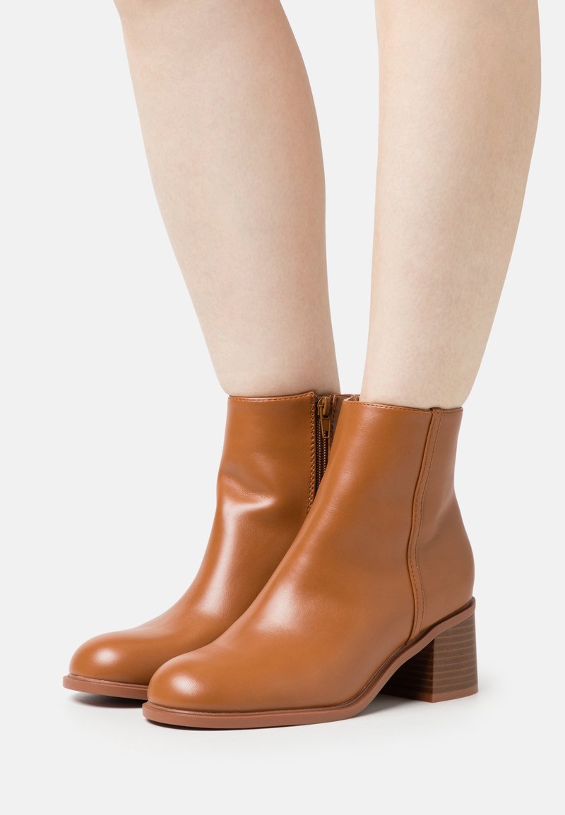 Bottes en cuir lisse couleur beige jusqu'à la cheville avec talons carrés, portées sur des jambes nues, montrées de face et de profil sur fond blanc.