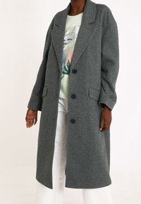 Lungo cappotto grigio a spina di pesce in un misto lana. Presenta un colletto a reverse, bottoni frontali e tasche laterali. Indossato sopra una t-shirt grafica verde chiaro.